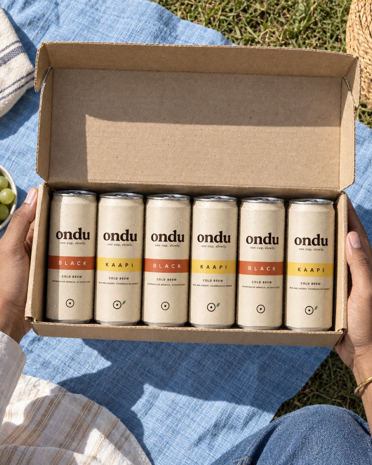 An open cardboard crate of mixed ondu cans held over a picnic blanket