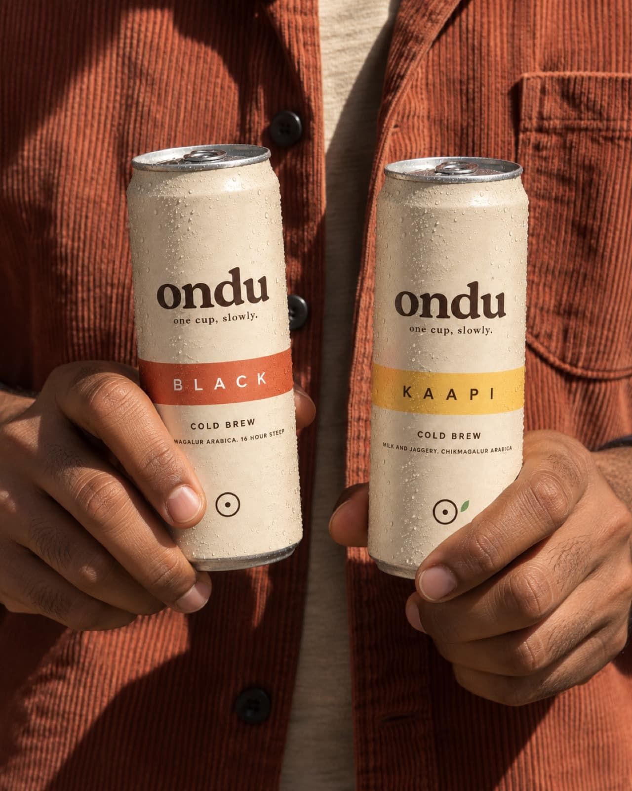 Two hands holding the BLACK and KAAPI ondu cans side by side